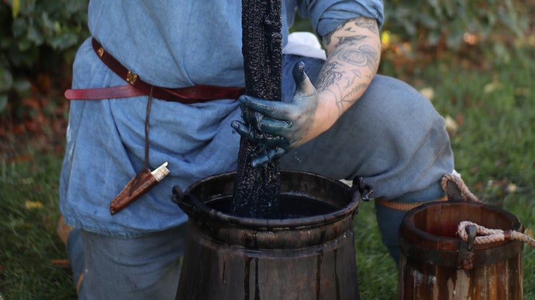 A pair of stained blue hands pulling a freshly-dyed skein of blue wool from a caldron